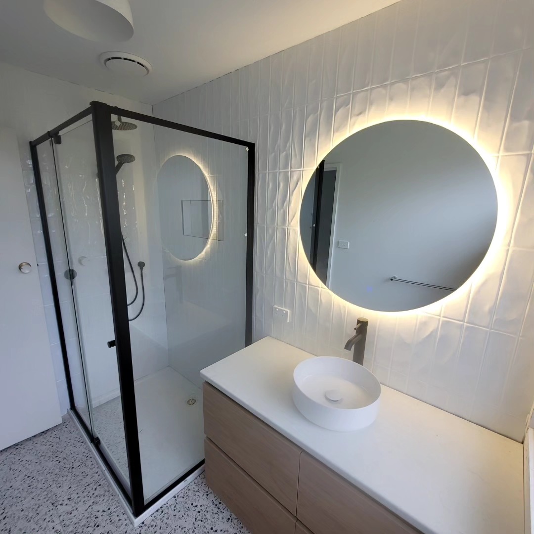 Renovated bathroom with a black-framed shower, backlit round mirror and timber vanity